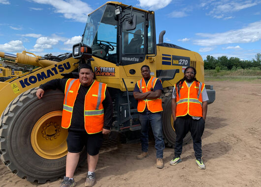 Three men in saftey vests standing in front of a wheel loader