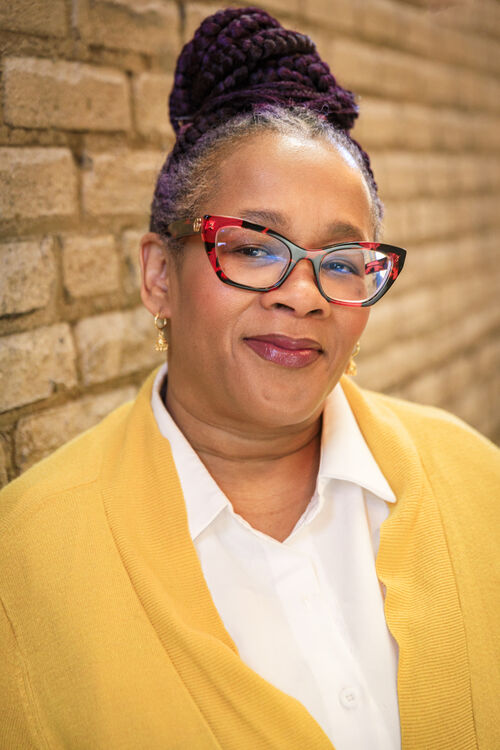 A headshot of Rashida Jackson wearing a yellow sweater standing against a brick wall 
